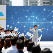 A group of people handling white flags and waving it over a political candidate standing and saying her speech on the stage. 